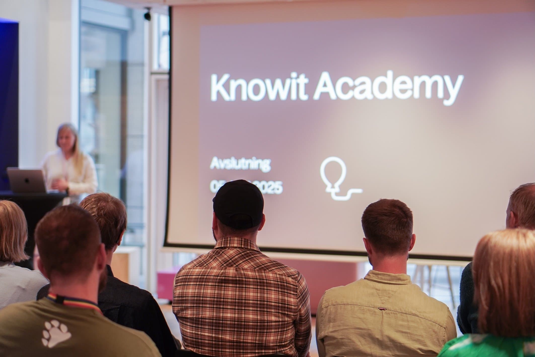 Mennesker som sitter foran en foredragsholder og en presentasjon på skjerm av Knowit Academy